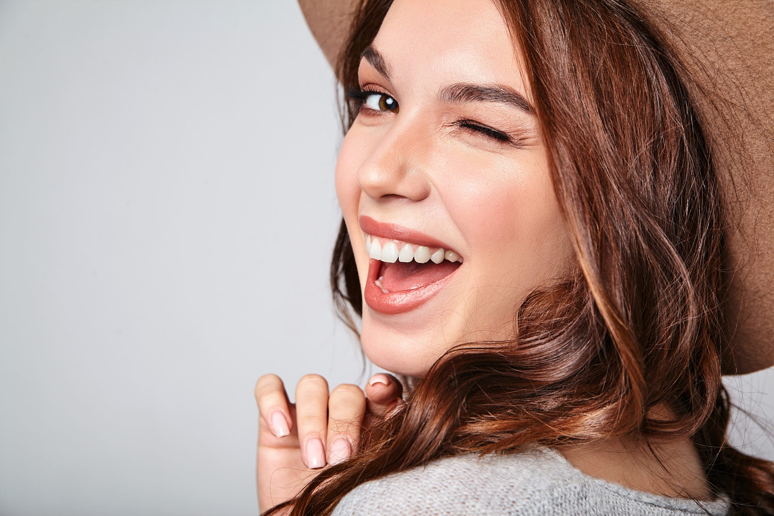 Portrait of young stylish laughing girl model in gray casual summer clothes in brown hat with natural makeup isolated on gray background. Looking at camera and winking chica joven con sombrero sonriendo y picando el ojo